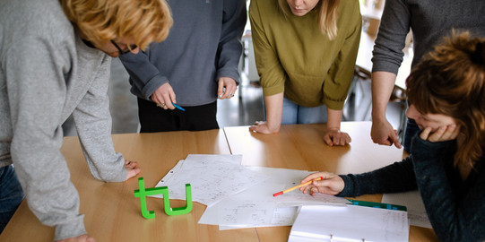 fünf Studierende stehen um einen Tisch und lesen ein Dokument.